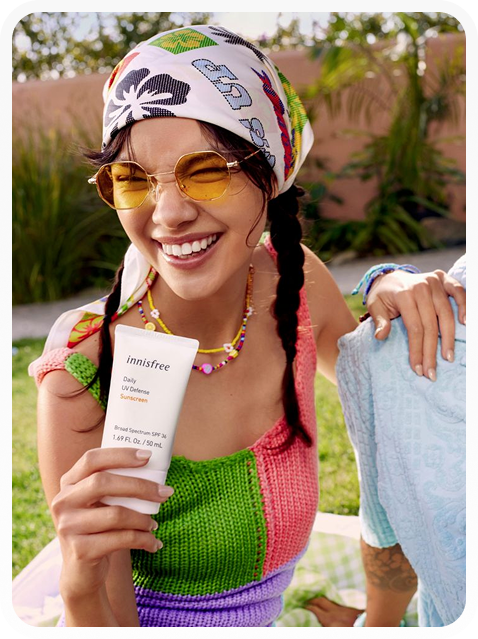 Smiling young woman wearing a colorful knitted top and a patterned headscarf holds a tube of Innisfree Daily UV Defense Sunscreen outdoors.